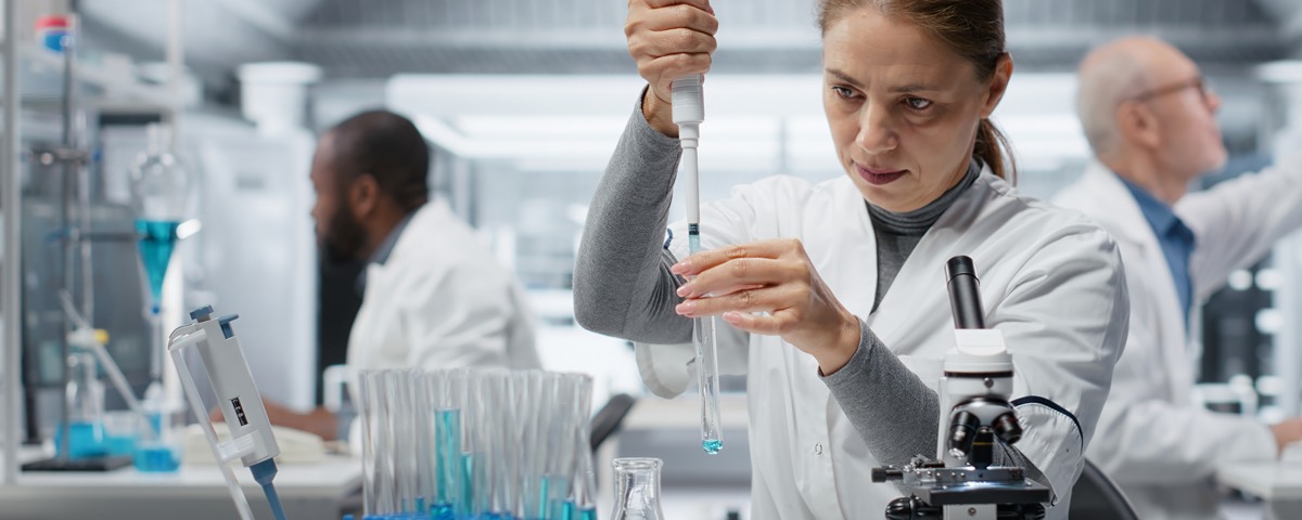 Scientist in white lab coat holding a pipette with a sample drop visible at the tip, with a microscope and sample racks containing blue solutions visible at the lab bench